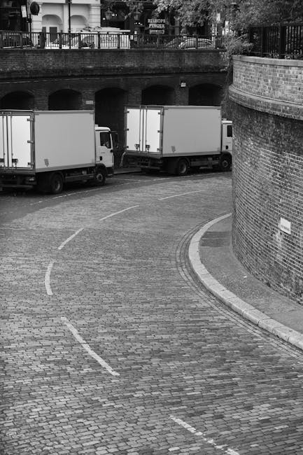 A black and white image showing two medium-sized box trucks with closed cargo doors parked side by side on a cobbled street near a brick wall and a curved sidewalk. The trucks are positioned adjacent to an opening under a bridge or viaduct, with a railing visible on top of the structure, suggesting an urban environment. The ground is paved with textured cobblestones, with white dashed lines marking parking or loading zones. The scene appears to be during daytime, with natural lighting illuminating the area, suitable for home relocation or furniture transport activities. This setting reflects the logistical aspects of house removals, with vehicles ready for loading or unloading near residential or commercial buildings, in line with services offered by Man with Van South Harrow regarding removals and moving logistics.