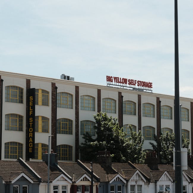 Exterior view of a large multi-storey building with a sign reading 'Big Yellow Self Storage' on the roof and a vertical black sign on the side that says 'Self Storage'. The building has numerous large, arched windows with yellow frames, and the facade features white walls with brick accents. In front of the building, there are several small residential houses with pitched roofs and chimneys. Two trees partially obscure some of the lower windows, and a streetlamp pole is visible on the right side of the image. The scene is lit with natural daylight under a partly cloudy sky, reflecting typical urban storage and relocation services environment, which aligns with house removals and furniture transport activities carried out by companies like Man with Van South Harrow.