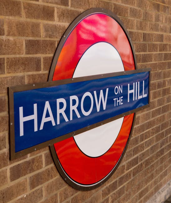 A close-up of a round London Underground sign mounted on a brick wall, featuring a red circle with a white center and a horizontal blue bar across the middle displaying the text 'HARROW ON THE HILL' in white letters. The sign is situated inside a building or station, with visible texture of the red brick wall behind it. The lighting appears even, illuminating the sign clearly. This image relates to the context of house removals and moving services by highlighting local area signage, which may be useful for clients navigating South Harrow during home relocation or furniture transport projects, with the sign indicating the Harrow-on-the-Hill area that can be relevant for service planning.