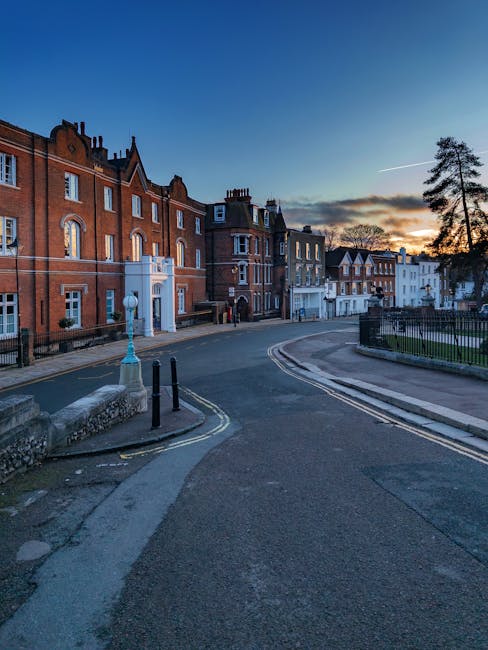 External view of a quiet street in South Harrow at sunset, lined with historic red-brick and white terraced houses with decorative brickwork and pitched roofs. A group of movers from Man with Van South Harrow are loading various packed cardboard boxes, wrapped furniture, and household appliances into a van parked nearby, using trolleys and blankets for protection. The scene captures the home relocation process, with the van positioned close to the property entrance, and a few individuals handling the loading and unloading of items. The street features a lamp post and bollards along the pavement, with a small park or green space bordered by a black metal fence on the right side. The lighting highlights the architectural details of the buildings, emphasizing the logistical aspects of furniture transport and packing for house removals in the South Harrow area.