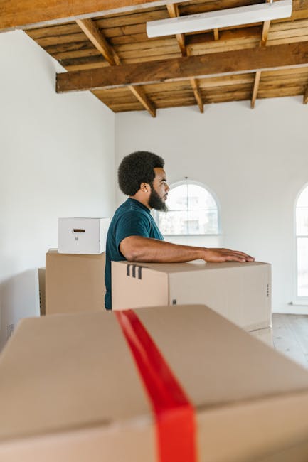 A man with a dark complexion, short curly black hair, and a beard, wearing a dark blue t-shirt, is inside a spacious room with a high wooden ceiling and two arched windows allowing natural light to enter. He is standing next to several cardboard boxes, with one box in front of him and others stacked behind him, some of which are sealed with red packing tape. The boxes vary in size and are likely used for packing belongings during a home relocation. The room has white walls and an unfinished wooden ceiling structure, indicating a possibly new or renovated property. The man is engaged in a packing or furniture transport process, typical of house removals carried out by services such as Man with Van South Harrow. There are no visible furnishings or appliances inside, and the scene captures the preparation aspect of a move, highlighting the handling and organization of packing materials in a home environment for a furniture transport or moving operation.