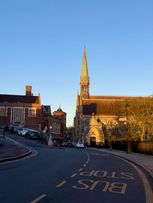 A quiet residential street in South Harrow during dusk, featuring a large white multi-story house on the left with a small front garden, a black metal balcony, and multiple windows. The street slopes 