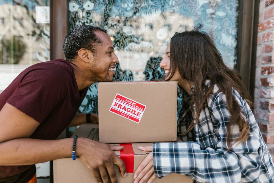 A man and woman engage in a home relocation exchange outdoors near a brick building with a window decorated with a patterned curtain. The man, with short curly hair and wearing a maroon T-shirt, leans forward smiling at the woman while holding a cardboard box labeled 'fragile' with red and white warning tape. The woman, with long dark hair, is dressed in a blue and white checkered shirt and smiles back, holding another cardboard box wrapped with red tape. Between them are two stacked cardboard boxes, one of which is being handed over or received, indicating a packing and moving process. Behind them, a van with an open door is visible, showing a glimpse of the interior space suitable for furniture transport and removals. This scene illustrates a professional moving service, such as Man with Van South Harrow, involved in logistical aspects of house removals and packing, emphasizing friendly cooperation during a local move.