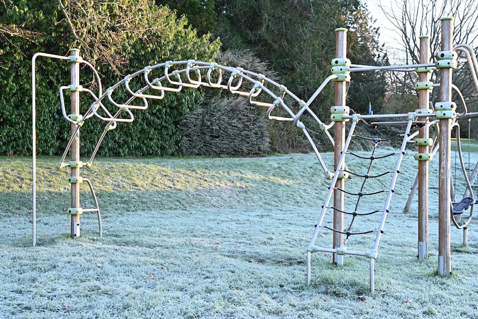 A frosty early morning scene at Roxeth Recreation Ground showing a children's climbing frame made of wooden posts, metal bars, and ropes, set on grass covered with thin frost. The climbing structure includes a curved horizontal ladder with rungs, a vertical climbing net with thick black ropes, and a small slide with a metal frame, all designed for children's play. In the background, dense bushes and tall trees are visible, with some branches bare, indicating winter conditions. The area appears calm and quiet with no children or people present. This outdoor environment is part of a park in South Harrow, and the image is relevant for illustrating outdoor play equipment that might be included in house removal or relocation services provided by Man with Van South Harrow, especially as part of the process of packing or preparing outdoor furniture and equipment for transport during a home relocation or furniture transport project.