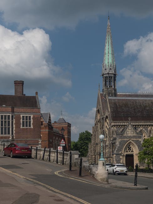 A street scene in South Harrow featuring a traditional stone church with a tall, pointed spire covered in greenish patina, situated next to a brick building with multiple windows and a chimney. The church has arched windows and detailed stonework above the entrance. In the foreground, there is a sloped pavement with a black metal railing along its edge, and a red car parked partially facing the church. A white car is parked near the church entrance, adjacent to a lamppost with a globe-shaped light. The sky is partly cloudy with patches of blue, casting natural daylight over the scene, which near [COMPANY_NAME] would be relevant for context about moving logistics in the area.