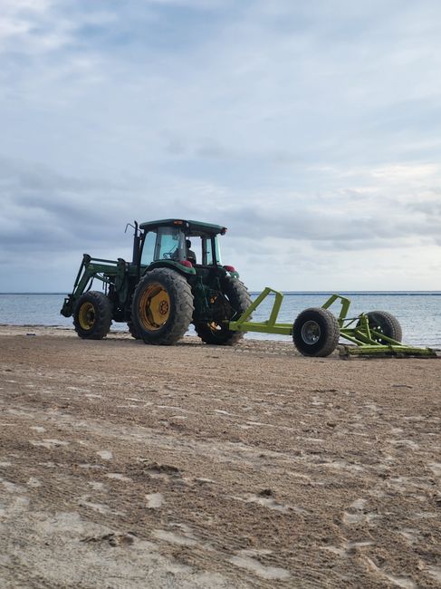 A green tractor equipped with large rear and smaller front tires is positioned on a sandy beach, with the ocean and a cloudy sky in the background. Attached to the tractor is a wide, green agricultural implement with two large wheels, which appears to be a piece of farming equipment or a soil aerator. The tractor is facing toward the sea, and the scene is illuminated by natural daylight, with no other vehicles or objects visible in the immediate vicinity. This image, while primarily showing machinery, could conceptually relate to the movement of heavy or bulky equipment during a home or commercial relocation, similar to furniture transport or loading processes in house removals handled by companies like Man with Van South Harrow.