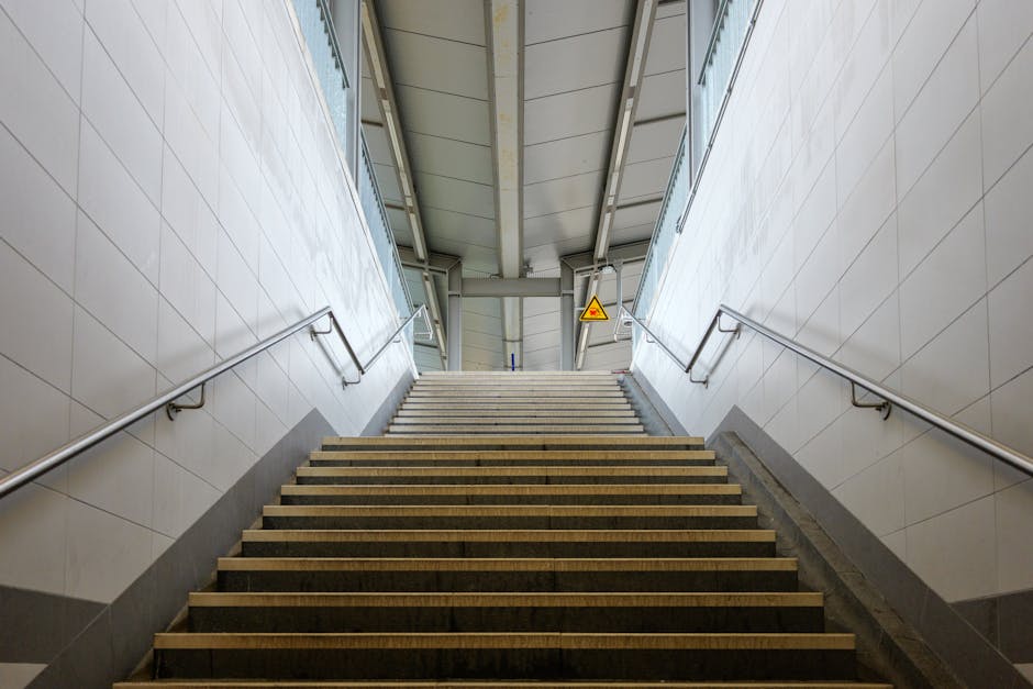 A symmetrical view of a staircase exiting South Harrow station with wide steps made of dark material featuring lighter edges, and metal handrails on both sides. The stairs are enclosed by white tiled walls, leading upward towards an open outdoor area. Overhead, the station's metal roof structure is visible, supported by beams, with a small triangular warning sign attached to the ceiling for safety. The area is well-lit, possibly by natural daylight, and the scene depicts a clean and orderly environment suitable for home relocation or furniture transport activities. This setting illustrates the typical transit environment associated with moving or packing services provided by Man with Van South Harrow, aligning with the station removals guide for flats and stairs.