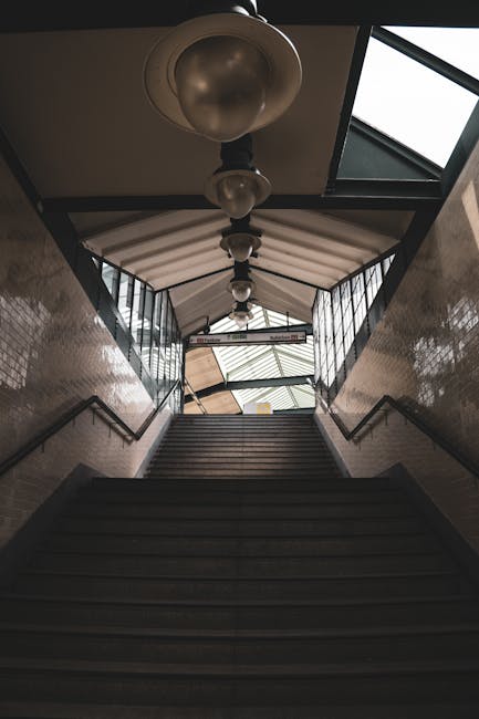 Inside a staircase corridor at South Harrow station, the image shows a downward view of a set of dark wooden steps leading to an open area with natural light flooding in through large glass windows at the top. The corridor is lined with beige tiled walls, with black metal handrails on each side. A series of vintage-style pendant lamps with frosted glass shades hang from the ceiling, spaced evenly along the hallway. Visible on the right side is part of a roof structure with a skylight, allowing additional daylight into the space. This setting illustrates a typical station interior, emphasizing the pathway used for pedestrian movement. In the context of house removals, furniture and boxes could be loaded or unloaded at such an entry point, supported by the presence of Man with Van South Harrow, a professional removals service specializing in home relocation and moving logistics. The image captures the process of transitioning between different environments, highlighting the importance of careful planning in furniture transport and packing during moves.