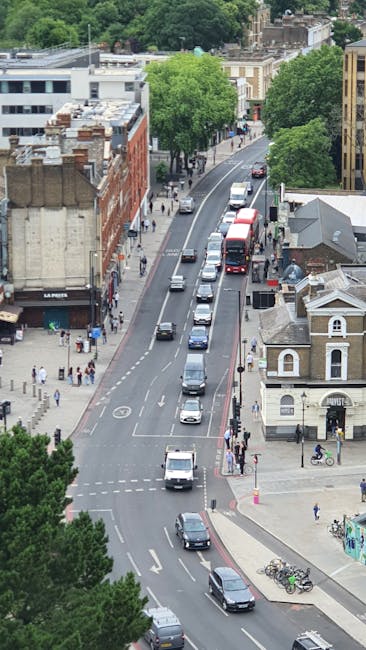 An aerial view of a busy urban street scene during daytime with parked and moving vehicles, including a red double-decker bus, cars, and vans. Sidewalks are populated with pedestrians, some waiting to cross and others walking along the pavement. There are several buildings lining the street, including a small white building with a dark roof and a larger multi-storey brick structure. Large, leafy green trees provide shade along the sidewalk. The street curves gently and extends into the distance amidst a backdrop of mixed residential and commercial properties, with roof terraces and chimneys visible on some buildings. The environment appears to be part of a town or city centre, and the photo captures the typical busy atmosphere of a home relocation or moving process in progress, with vehicles possibly waiting to load or unload furniture and packing materials, supported by local removals company Man with Van South Harrow.