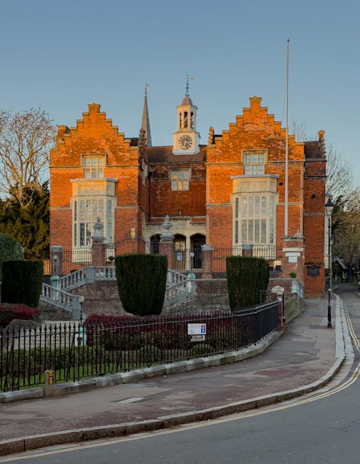 No visible interior in the image; the photograph shows a street scene during daylight, featuring a historic stone church with a tall, pointed spire and Gothic architectural details, situated on the right side. To the left, there are brick buildings with pitched roofs and multiple windows, some with decorative elements. The street has a curved, paved surface with yellow markings indicating a bus stop, and a line of parked cars along the roadside. The scene includes a clear blue sky with minimal clouds, and the area appears calm, likely indicating a residential or mixed-use neighbourhood, relevant to house removals and moving services offered by Man with Van South Harrow at [PAGE_TITLE], helping with home relocation and furniture transport through effective packing and loading processes.