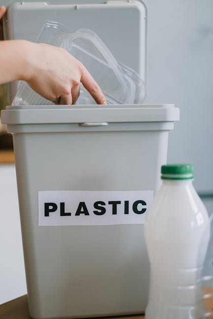 A person wearing a white shirt and beige trousers is holding a transparent plastic bottle with a red cap, preparing to dispose of it. The individual is standing next to a grey recycling bin labeled 'P